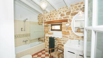 Un baño con pared de piedra, una bañera, un inodoro, una silla de madera y un espejo.