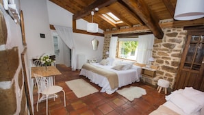 Un dormitorio rústico con pared de piedra, techo de madera, cama con ropa de cama blanca, un taburete de madera y una mesa con sillas.