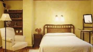 Un dormitorio con techo de madera, una cama con cubrecama blanco, una silla, una mesita de noche con lámpara y un estante con adornos.