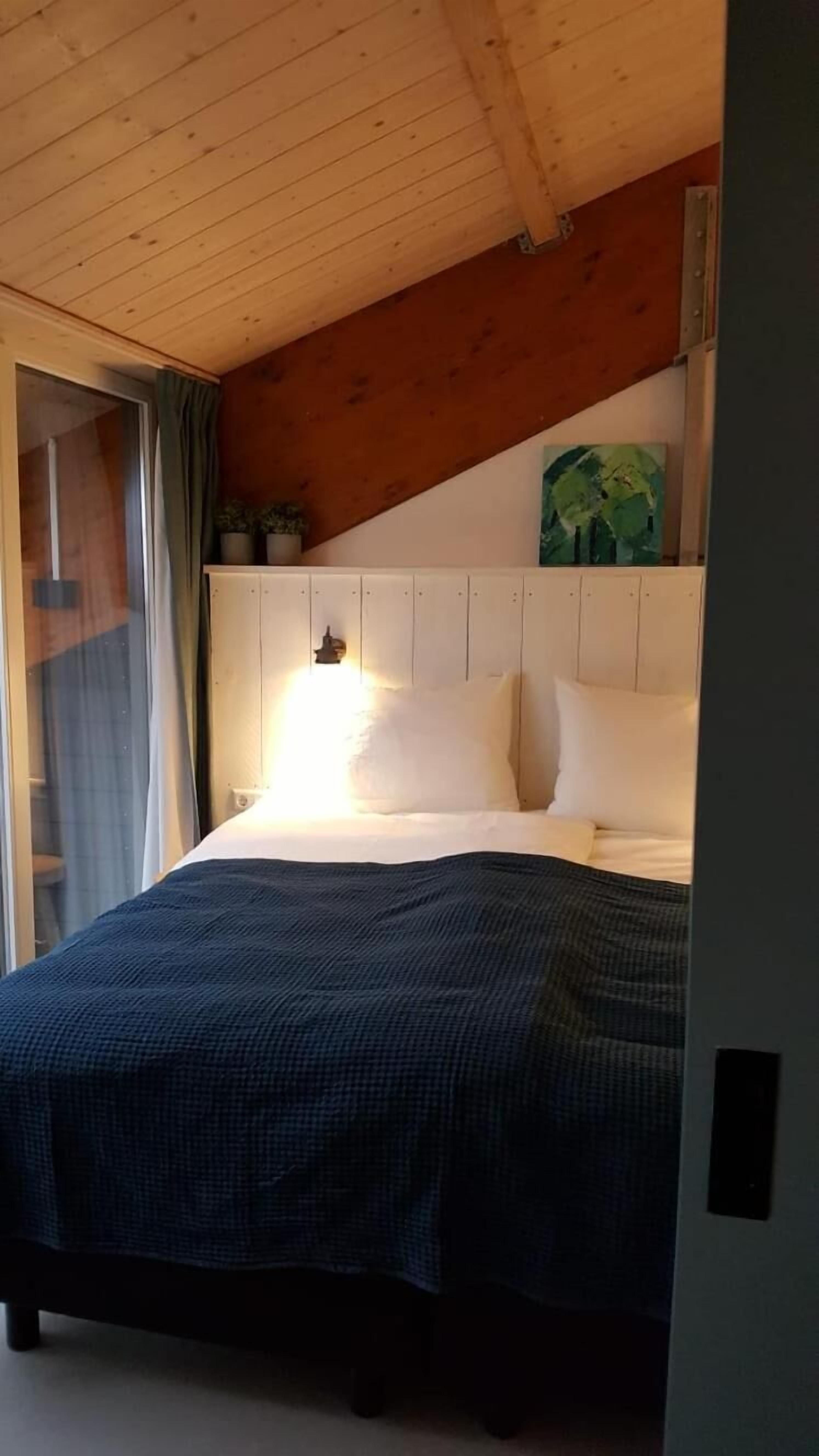 Un dormitorio con techo de madera, una cama con ropa de cama blanca, una manta verde y azul a cuadros y una planta verde en la pared.