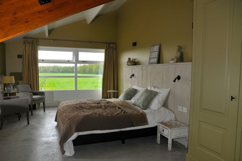 Un dormitorio con cama, mesita de noche y vistas a un paisaje verde a través de un ventanal grande.