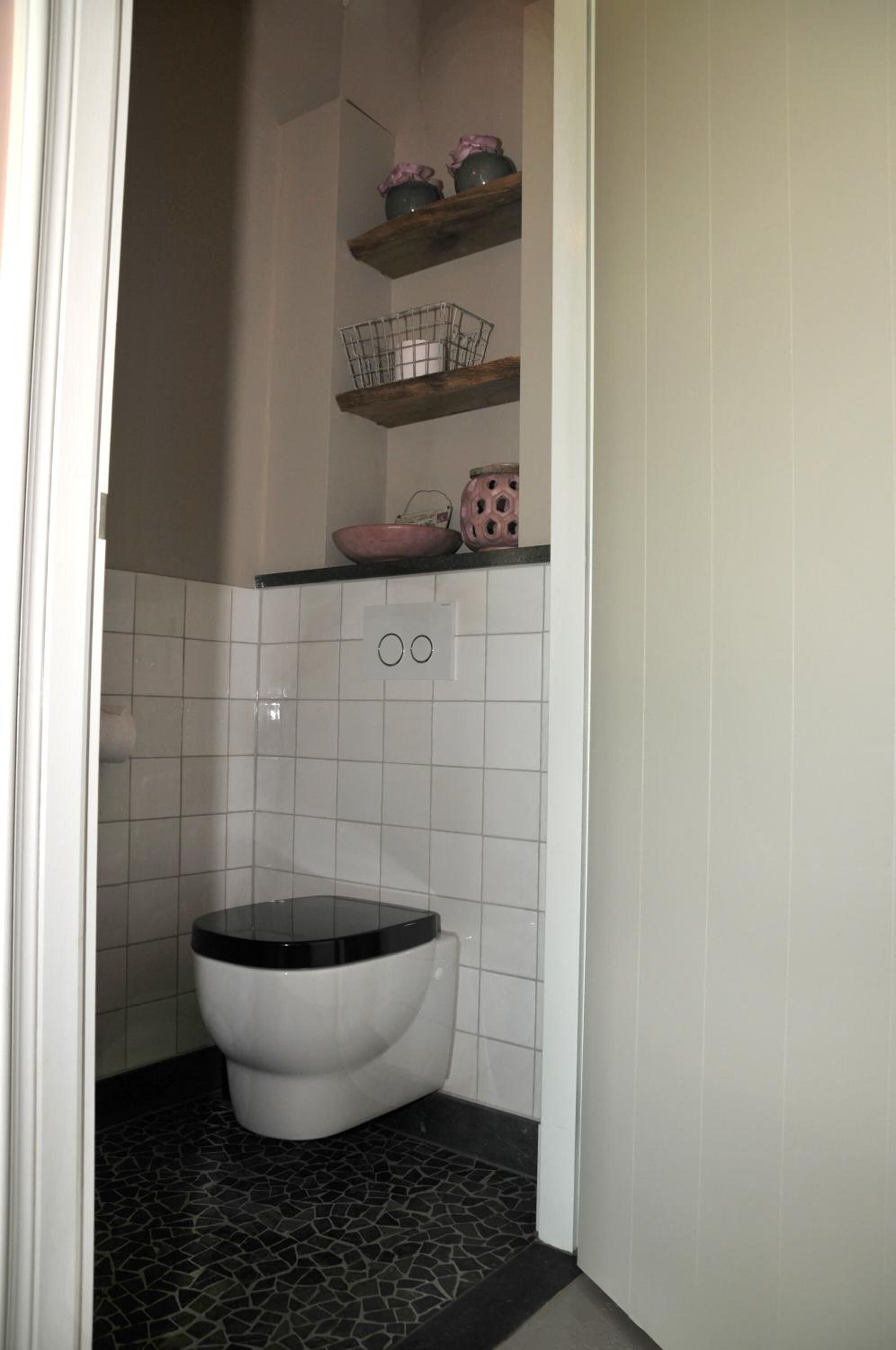 A small bathroom with a toilet, white tiled walls, a black tiled floor, and a shelf with decorative items.