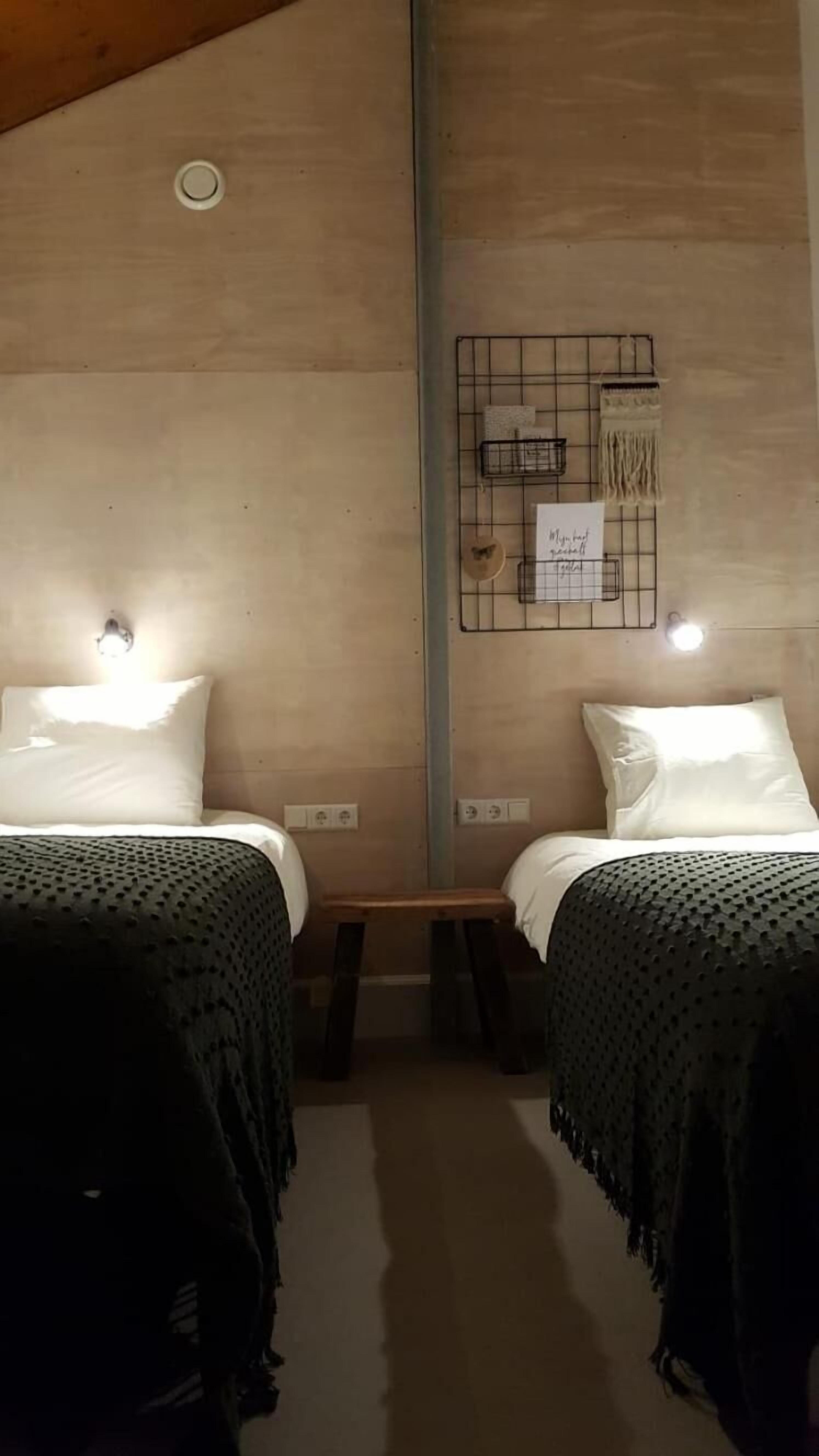 A room with two beds, each with a white pillow and a black textured blanket. There is a small wooden stool between the beds, and a wall-mounted shelf with a basket and a hanging item.