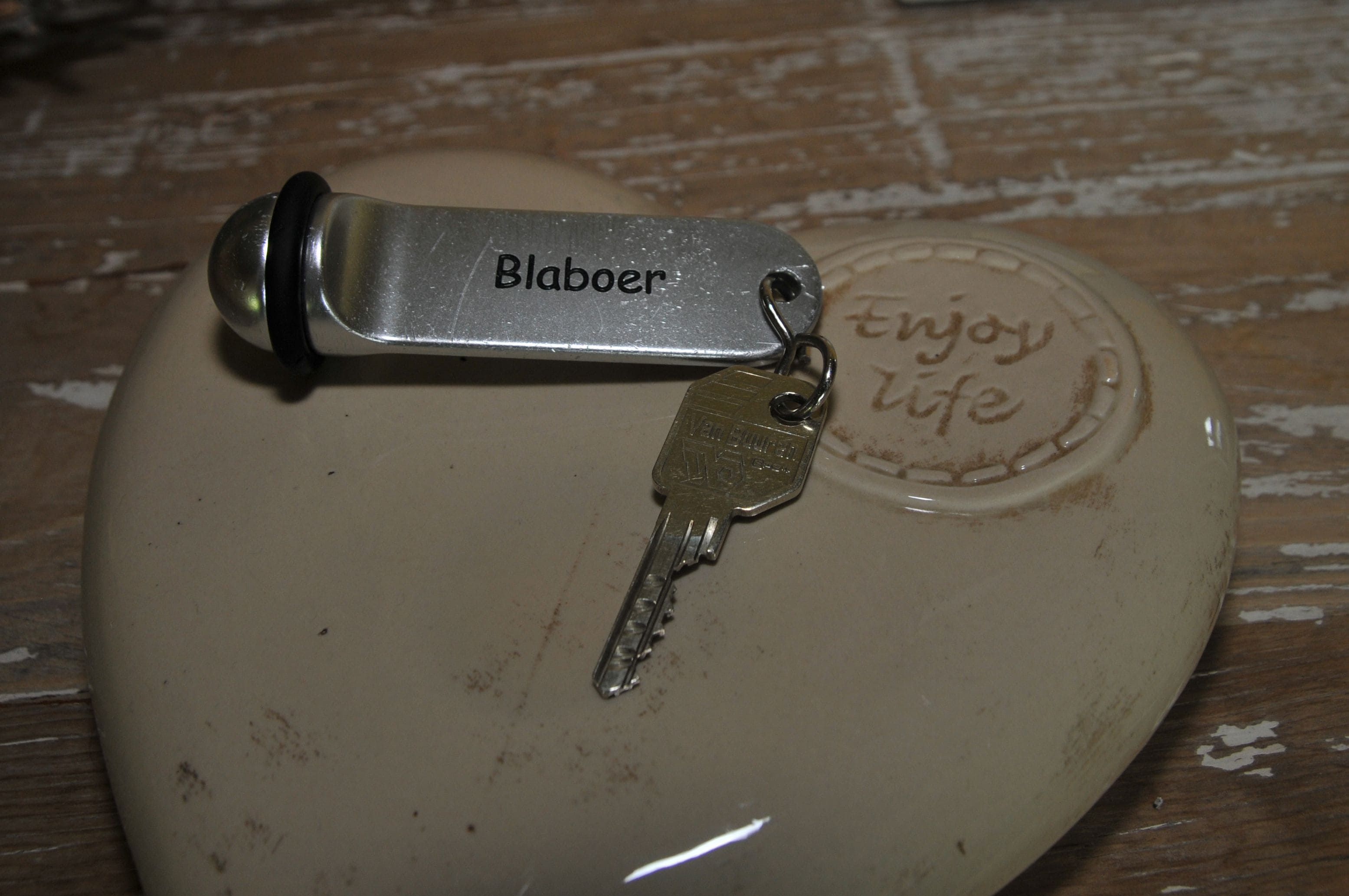 A key and a metal tag with the inscription "Blaboer" on a beige ceramic object with a "Enjoy life" engraving.