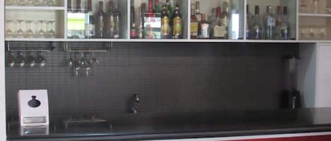 A bar area with shelves of bottles and glasses, a tiled backsplash, and three white bar stools.