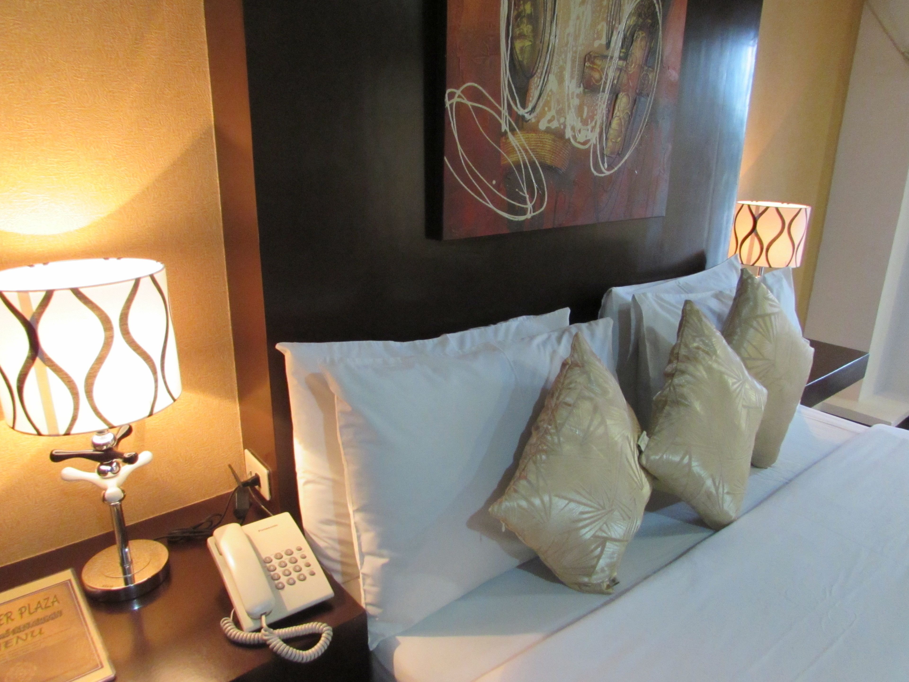 A hotel room with a bed, pillows, a bedside lamp, a phone, and a painting on the wall.