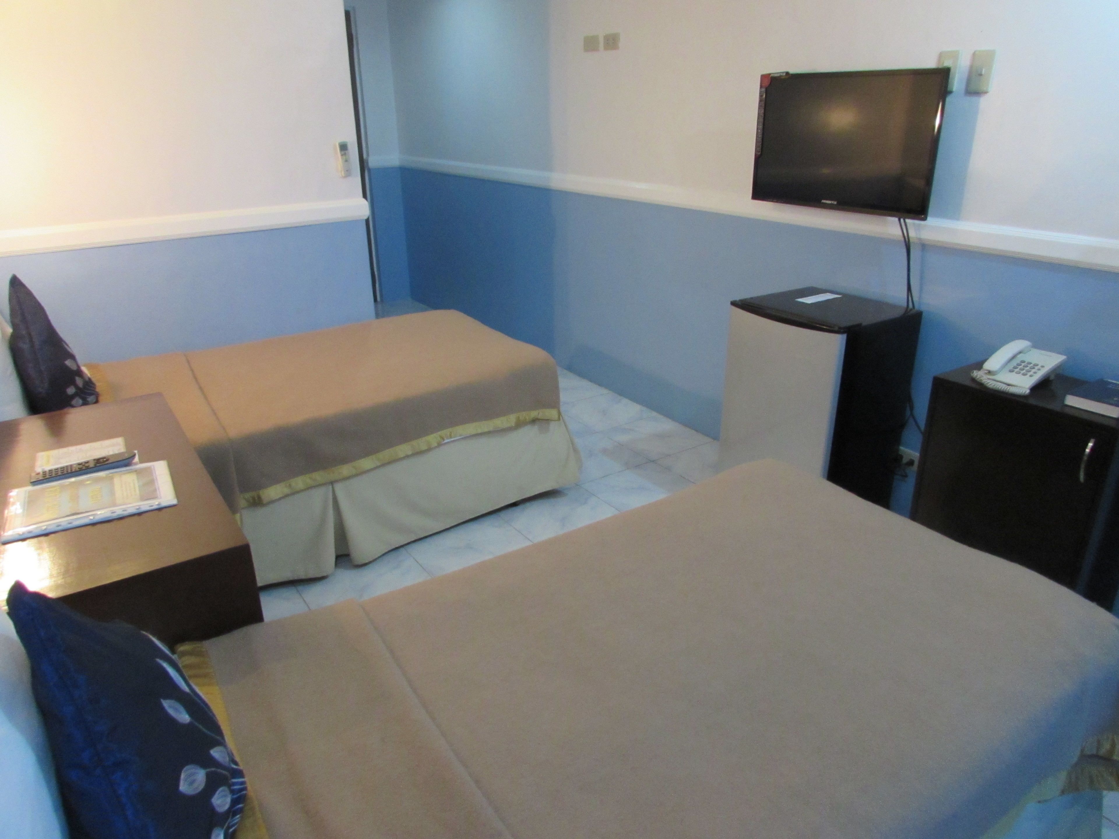 Une petite chambre d’hôtel, de taille modeste, comprenant un lit simple, un bureau avec un téléphone, un téléviseur fixé au mur et un petit placard.