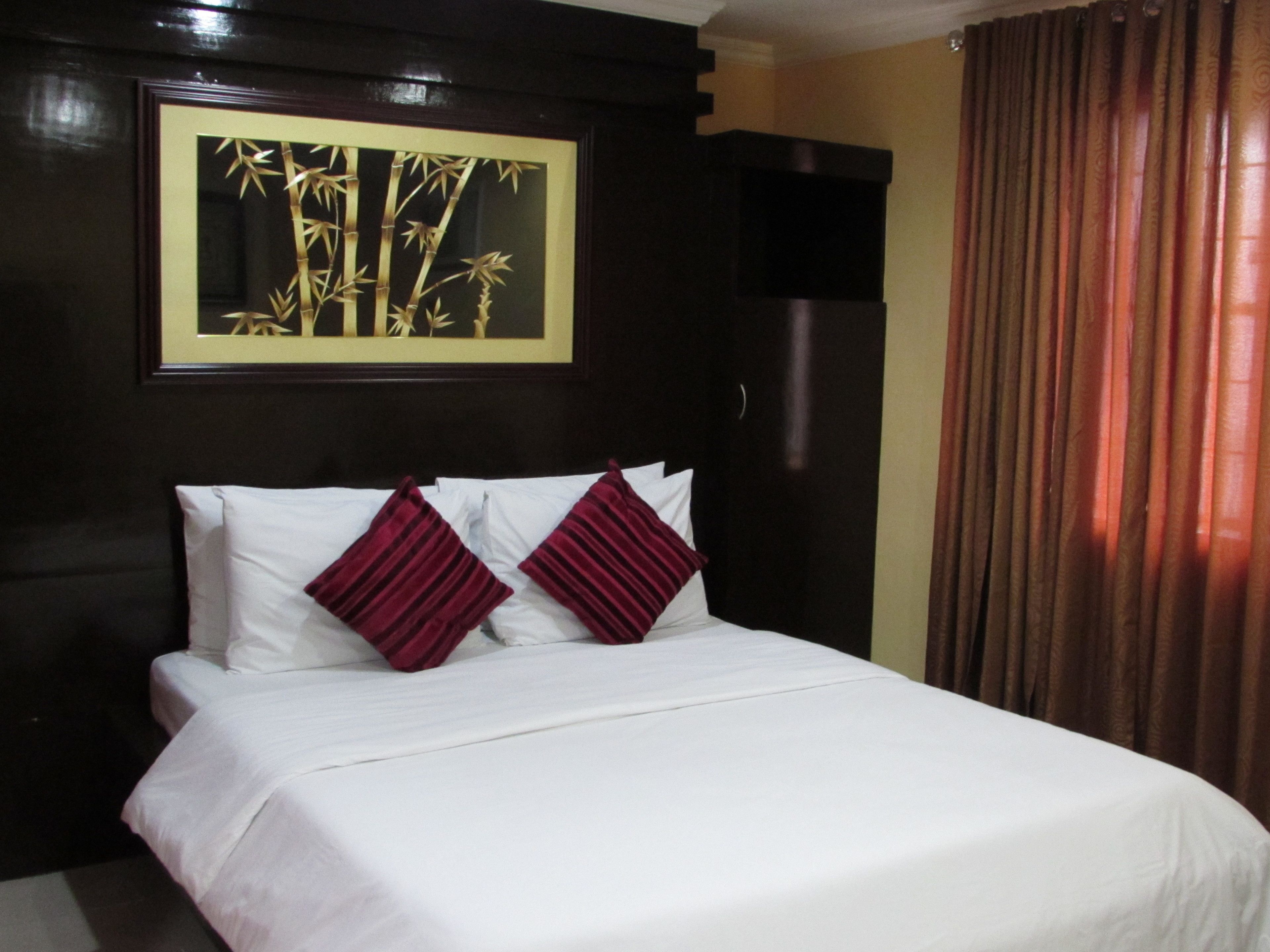 A neatly made bed with white linens and two red pillows, a framed artwork of bamboo on the wall, and a dark wooden headboard.