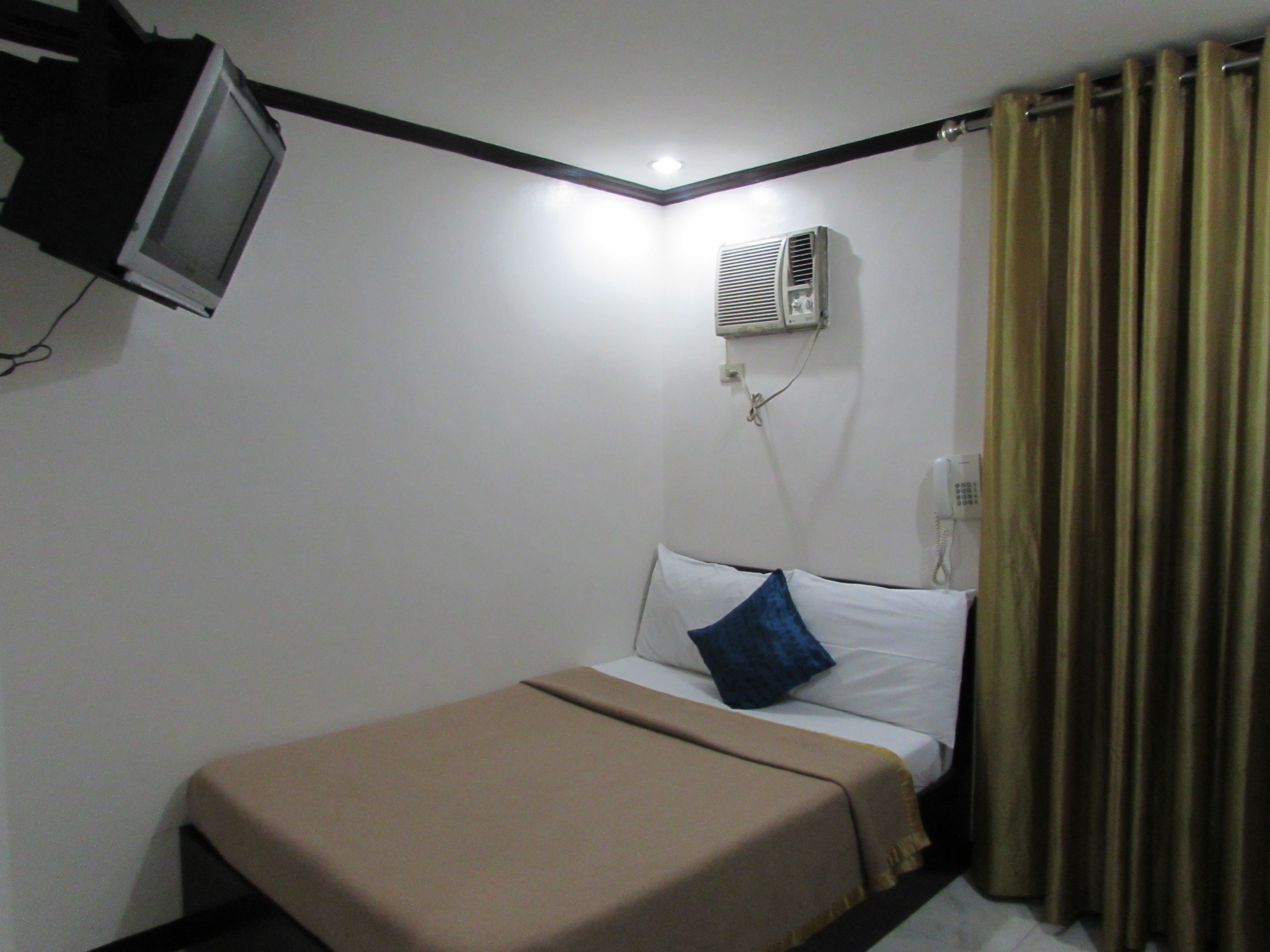 A small, single-bed room with a television mounted on the wall, an air conditioning unit, a telephone, and a curtain.