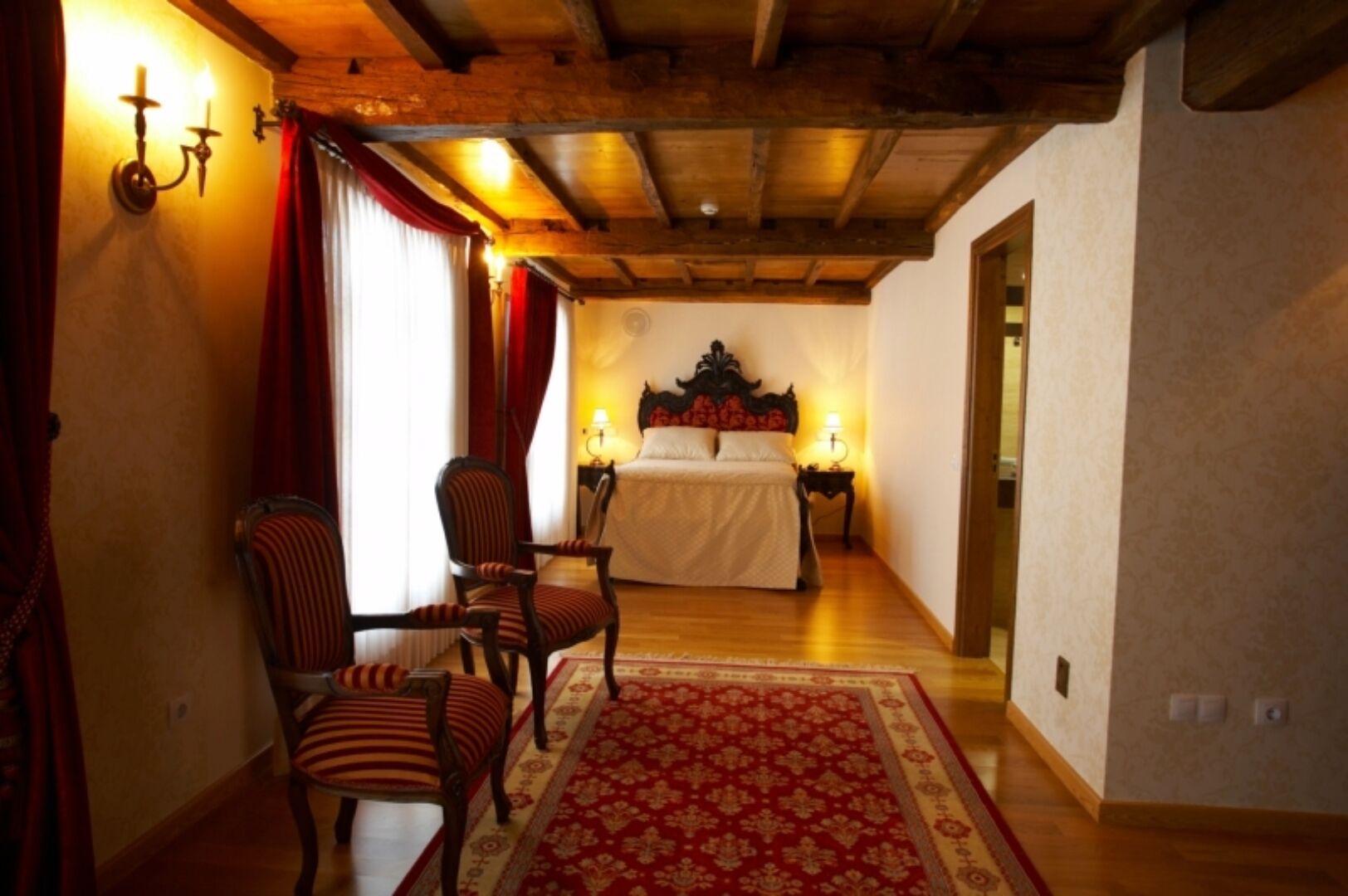 Een slaapkamer met een houten plafond, een bed met een witte dekbedovertrek, een gestreepte stoel in rood en wit, en een tapijt met een rood patroon.