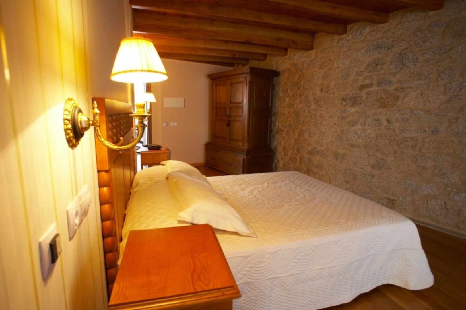 Un dormitorio con cama de madera, mesita de noche, lámpara y pared de piedra.