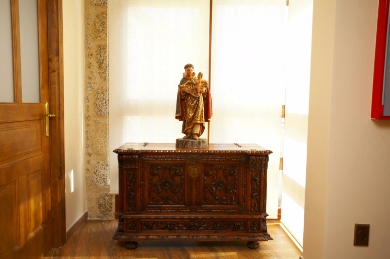 A wooden cabinet with a statue on top, located in a hallway with a wooden door and patterned wallpaper.