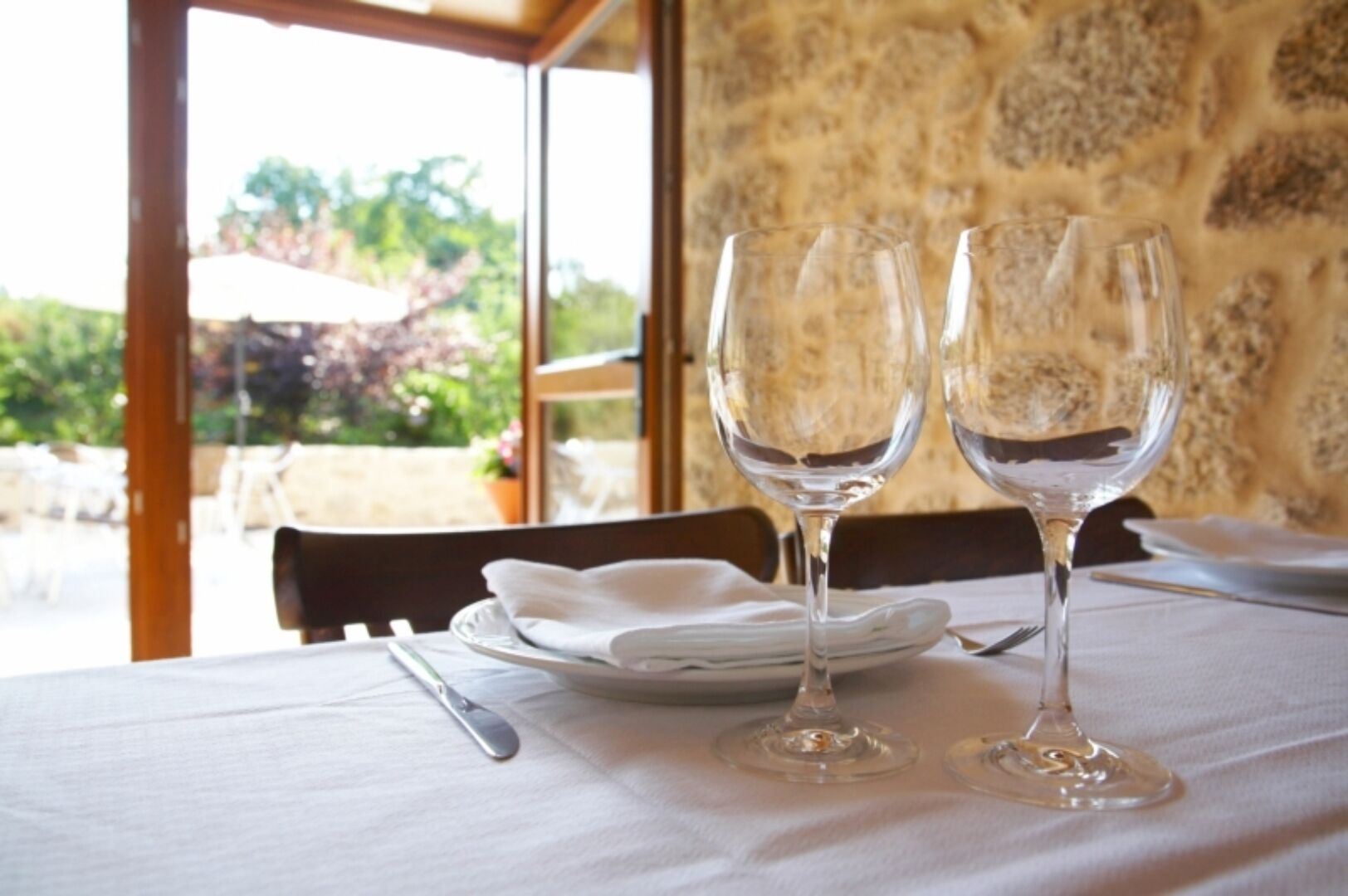 A table set with two wine glasses, a fork, and a plate, with a view of greenery outside.