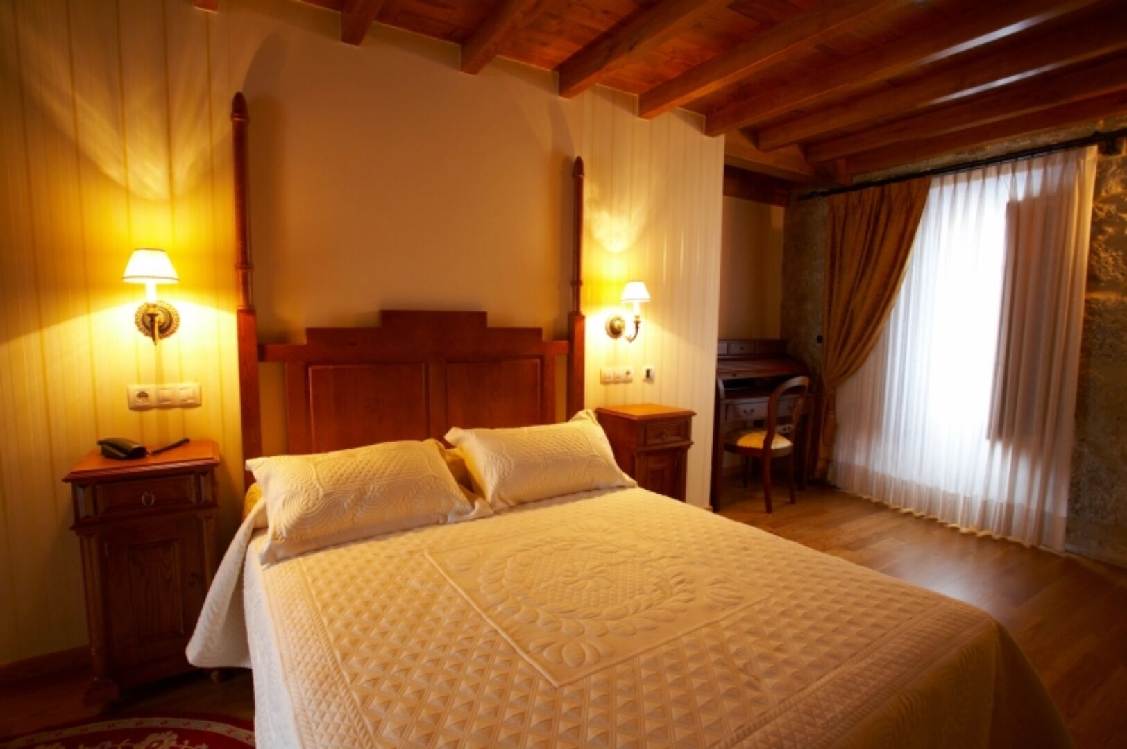 A hotel room with a large bed, wooden headboard, two bedside tables with lamps, a desk, and a chair.