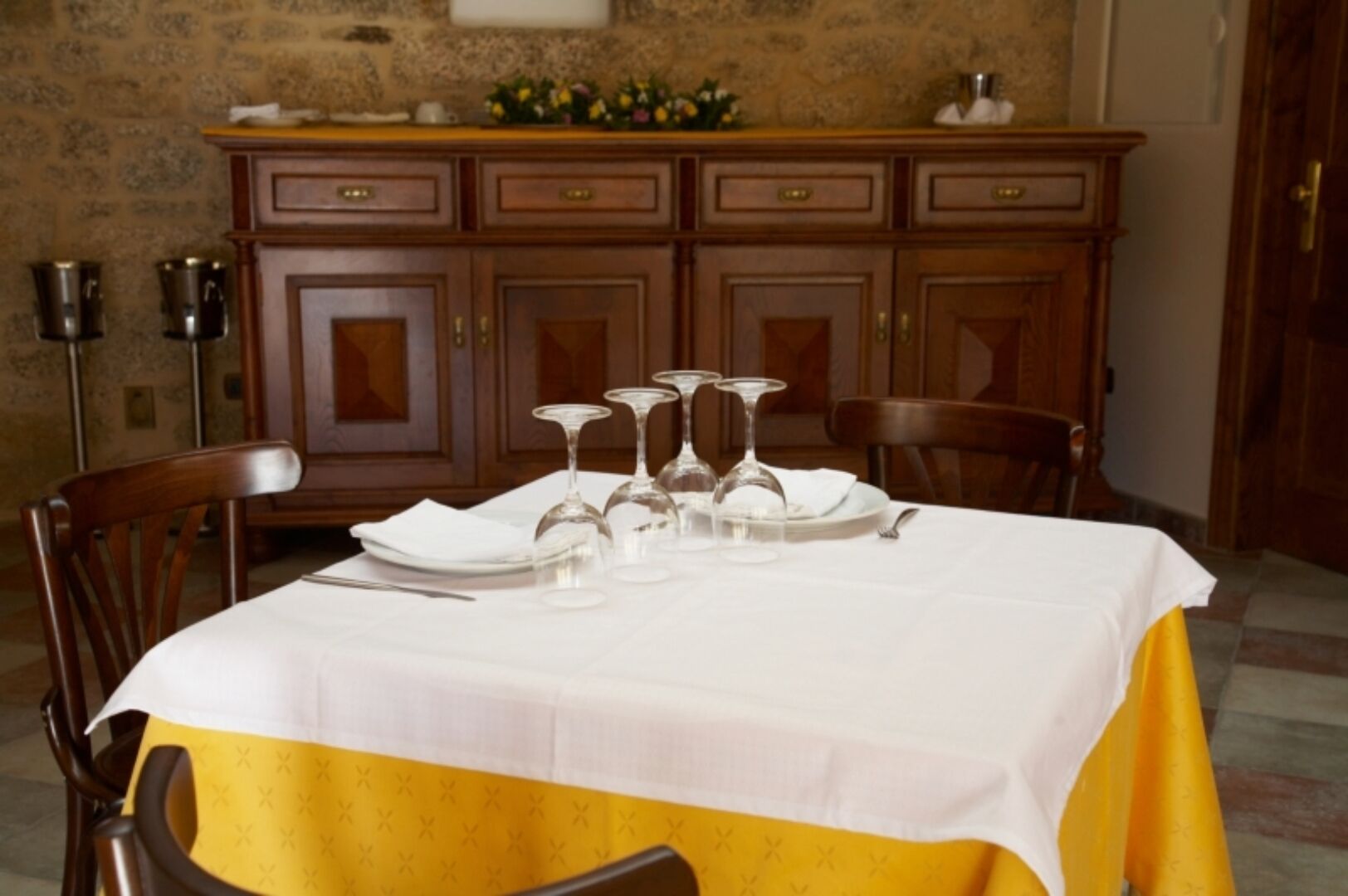 A dining table set with white tablecloth and yellow accents, flanked by wooden chairs.