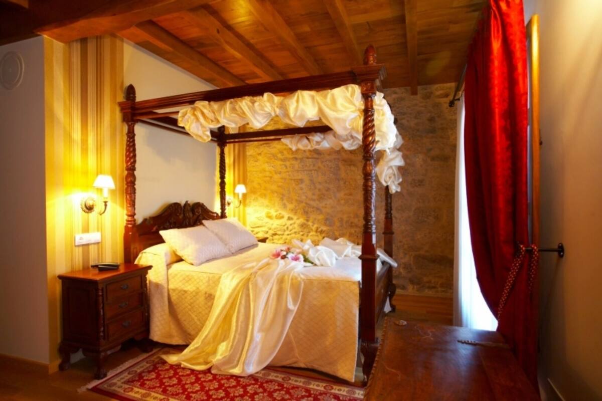 A four-poster bed with a canopy, a wooden nightstand, a red patterned rug, and a stone wall in the background.