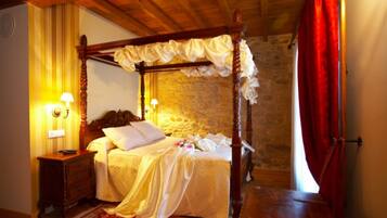 Una cama con dosel, un mesita de noche de madera, un tapete rojo estampado y una pared de piedra al fondo.