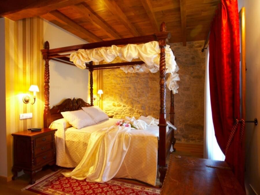 Una cama con dosel, un mesita de noche de madera, un tapete rojo estampado y una pared de piedra al fondo.