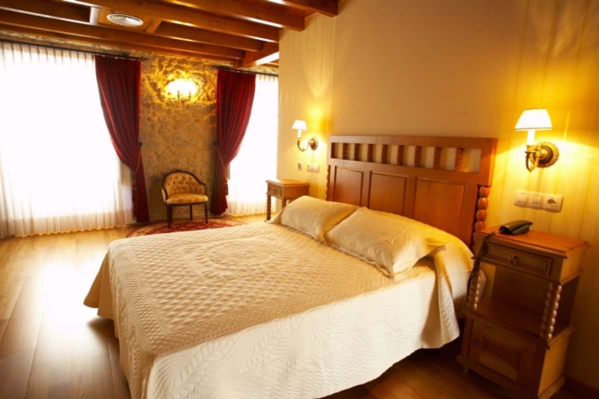 A hotel room with a large bed, wooden headboard, two bedside lamps, a chair, and a stone wall.