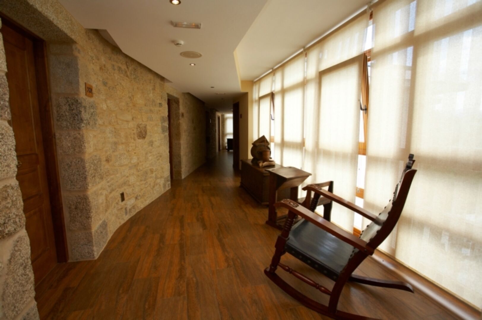 Un pasillo con paredes de piedra, suelo de madera y una mecedora cerca de una ventana con cortinas translúcidas.