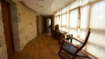 Un pasillo con paredes de piedra, suelo de madera y una mecedora cerca de una ventana con cortinas translúcidas.