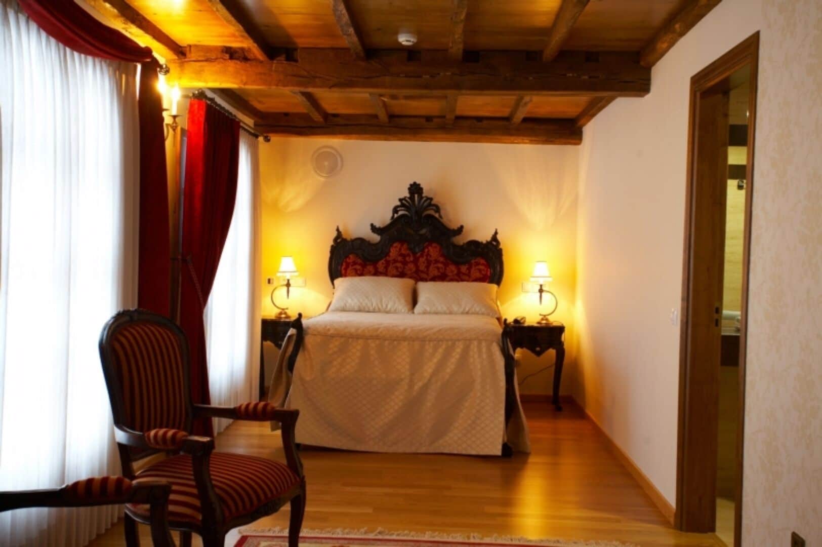 Une chambre avec un plafond en bois, un grand lit avec une tête de lit, deux tables de chevet avec des lampes, une chaise et une fenêtre avec des rideaux.
