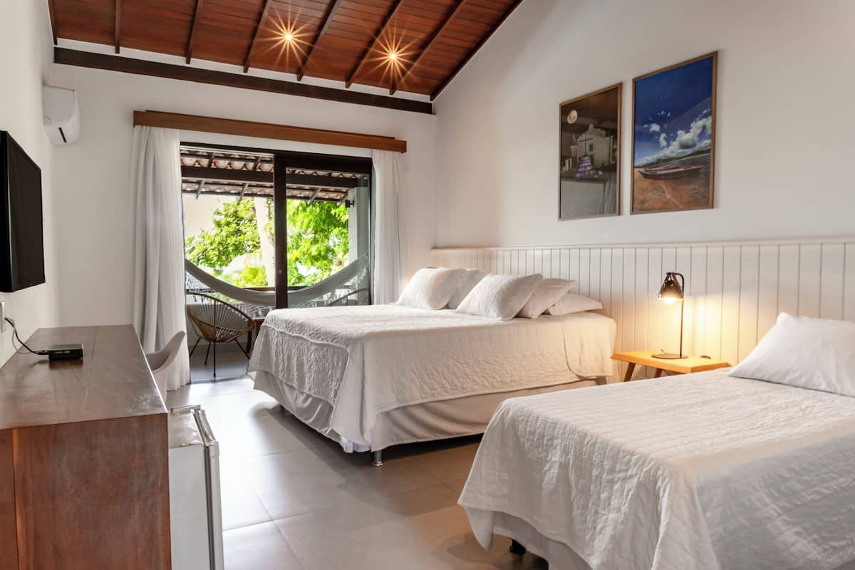 A hotel room with two beds, a desk, a chair, a hammock, and a view of greenery outside.