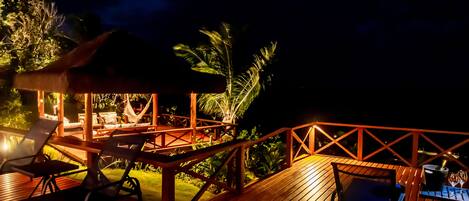 Une terrasse en bois, agrémentée de chaises longues et d’une table, entourée d’une végétation luxuriante et d’une structure à toit de chaume.