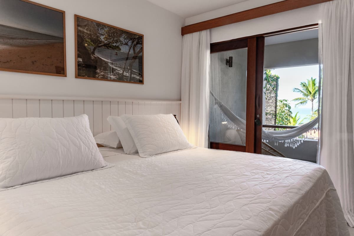A bedroom with a large bed, white bedding, a wooden headboard, two framed pictures, and a view of a hammock and palm trees through a glass door.
