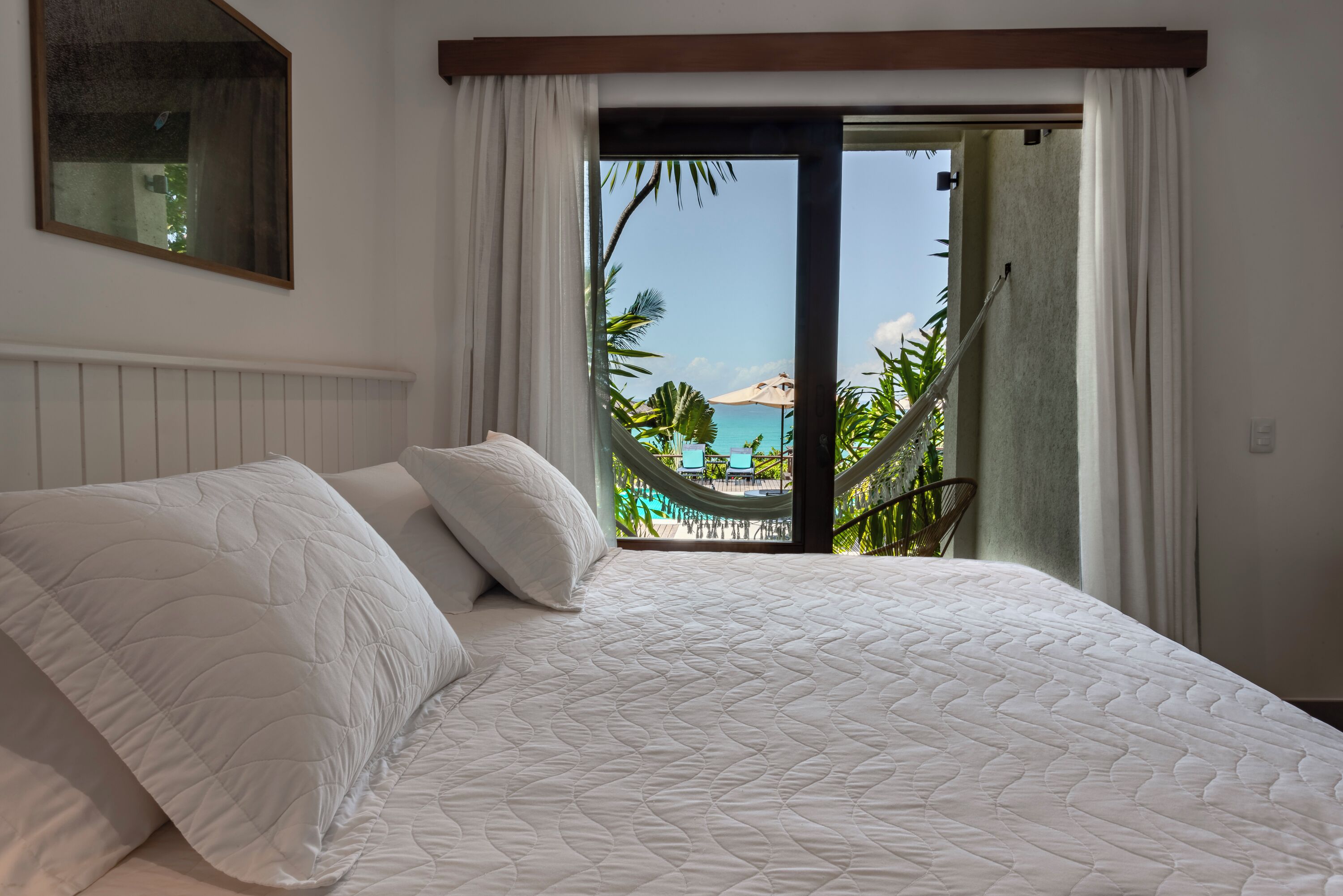 Quarto com cama grande, roupa de cama branca e vista para uma piscina e palmeiras através de uma porta de vidro.