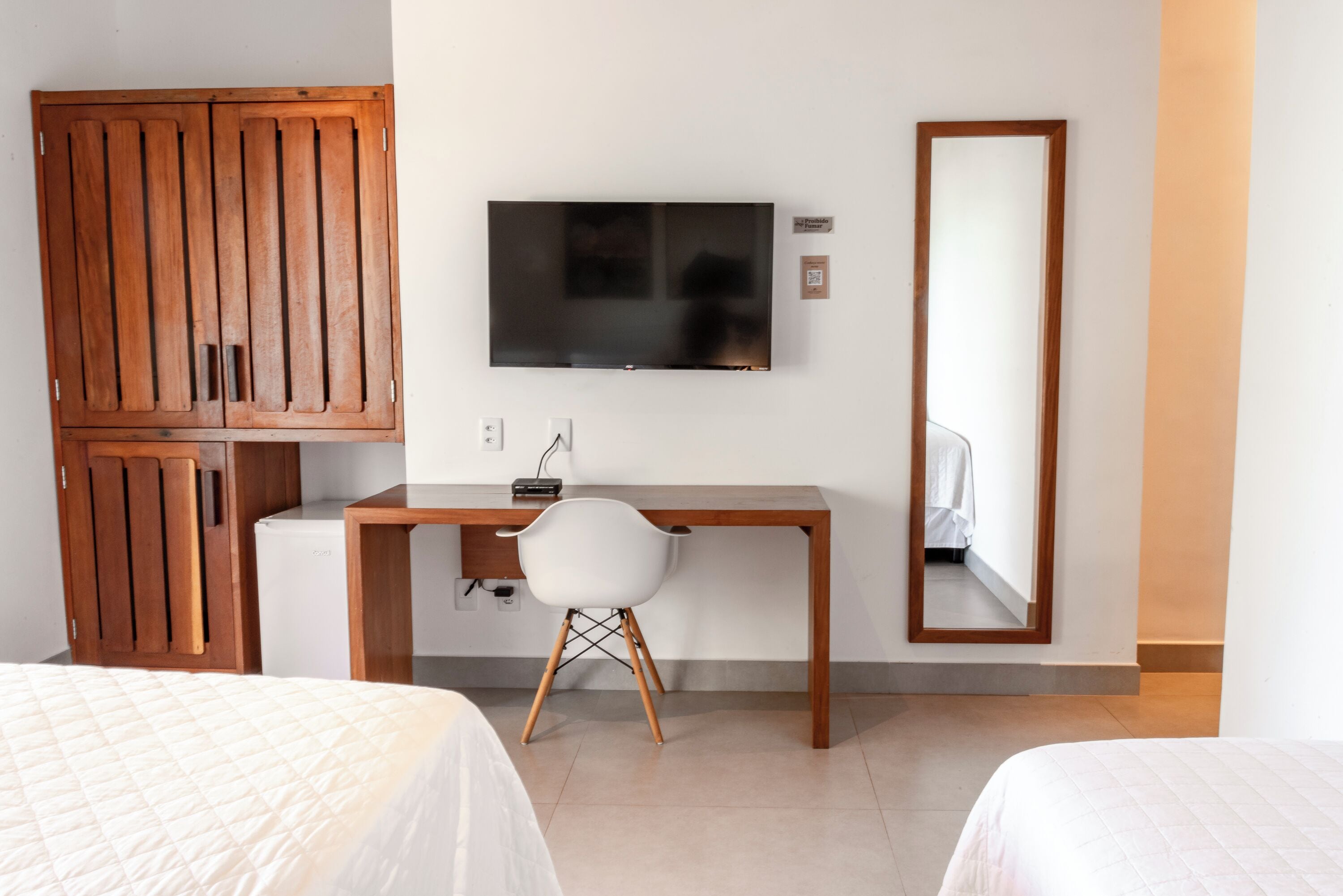 A hotel room with a wooden wardrobe, a desk with a chair, a TV mounted on the wall, and a mirror.