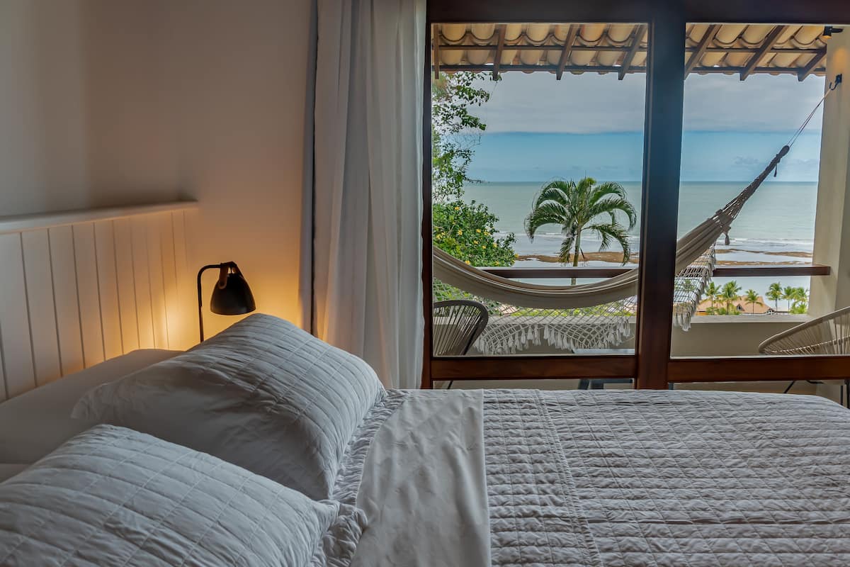 A bedroom with a view of the ocean, a hammock, and a bed with white bedding.