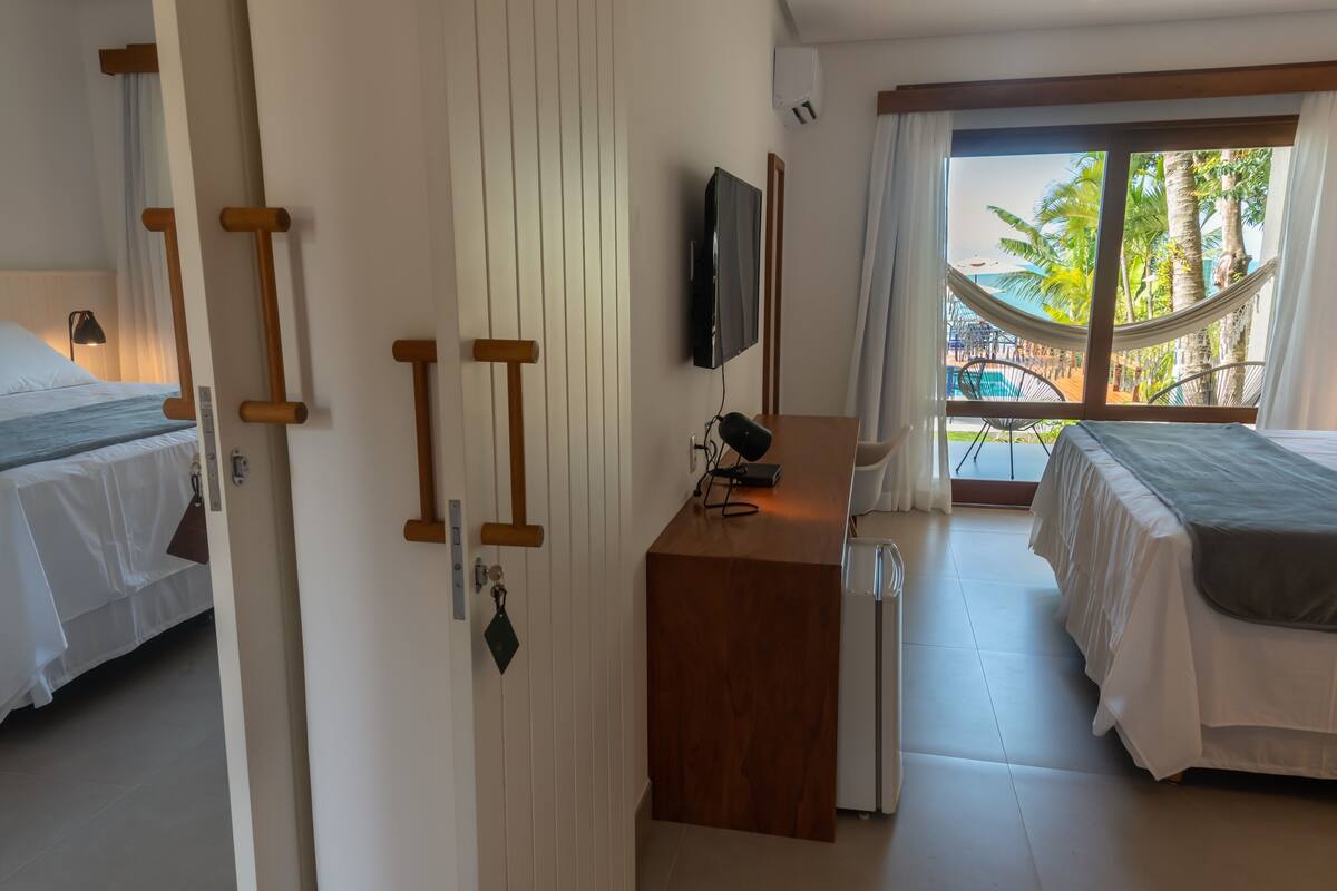 A hotel room with a bed, a desk, a TV, and a balcony with a view of palm trees.