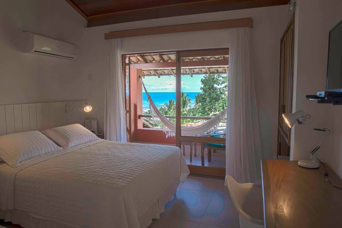 A bedroom with a bed, a desk, a chair, a window with a view of the ocean, and a hammock outside.
