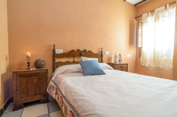 A bedroom with a wooden headboard, a white bedspread, a blue pillow, a nightstand with a candle, and a window with curtains.