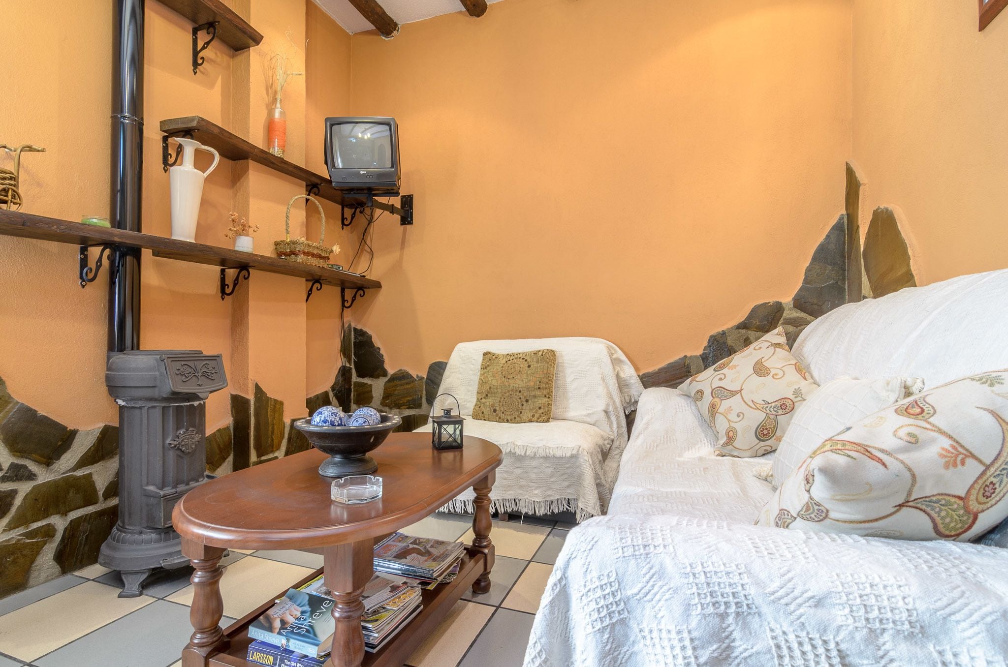 A cozy living room with a stone wall, a fireplace, a white sofa with patterned cushions, a wooden table with magazines, and a mounted television.