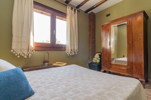 A bedroom with a bed, a wooden wardrobe, a window with curtains, and a small table with a basket and a hat.