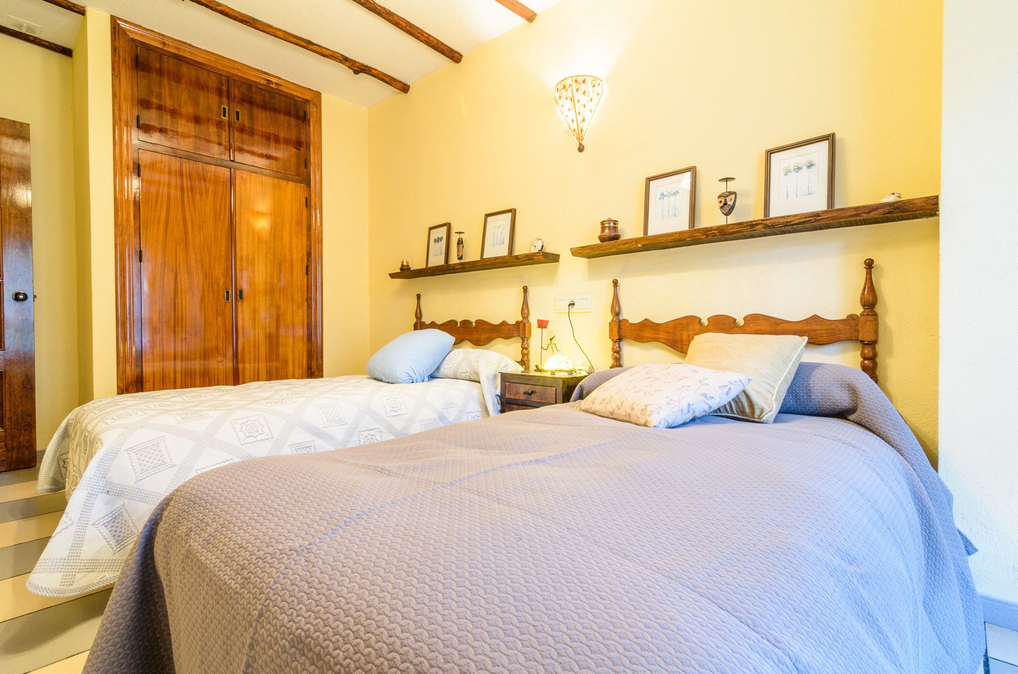 A bedroom with two beds, wooden wardrobe, wall-mounted shelves with decorative items, and a chandelier.