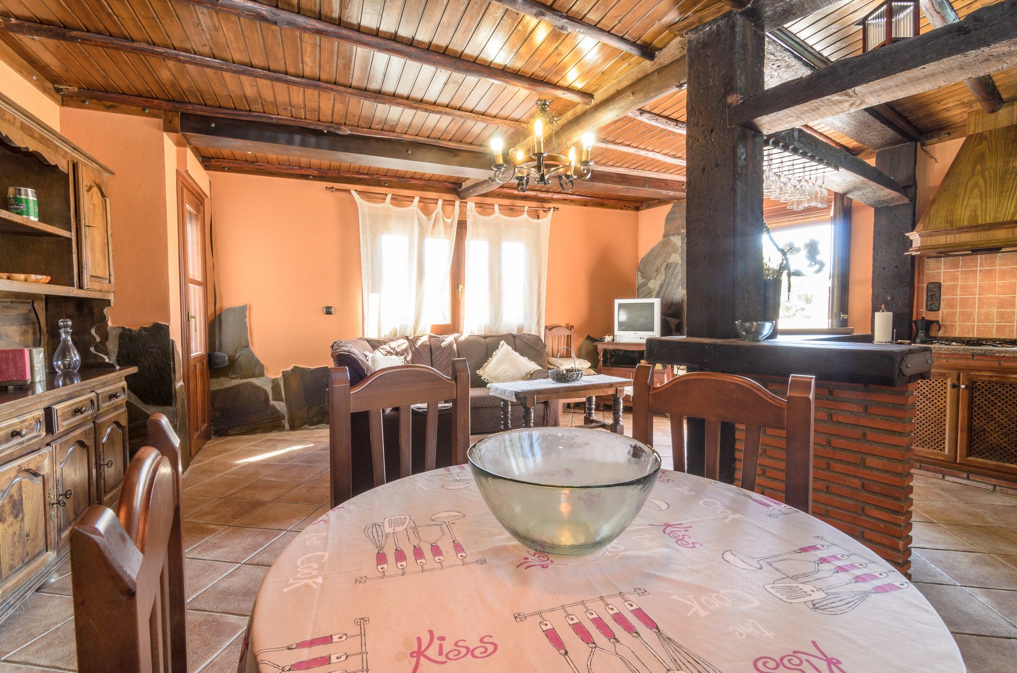 A cozy kitchen with wooden beams, a dining table with a bowl, and a stone fireplace.