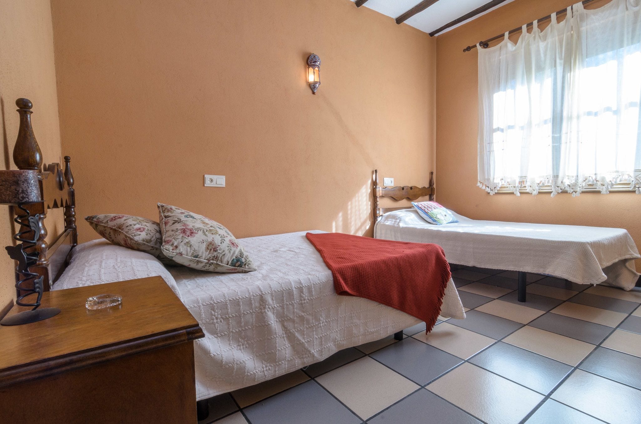 Une chambre avec deux lits, une table de chevet en bois, une lampe et une fenêtre avec des rideaux.