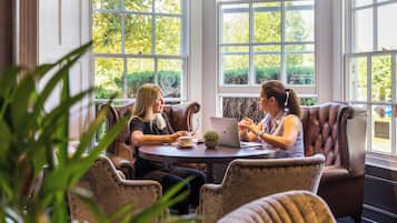 Twee personen zitten aan een ronde tafel met een laptop, in een kamer met grote ramen en lederen stoelen.