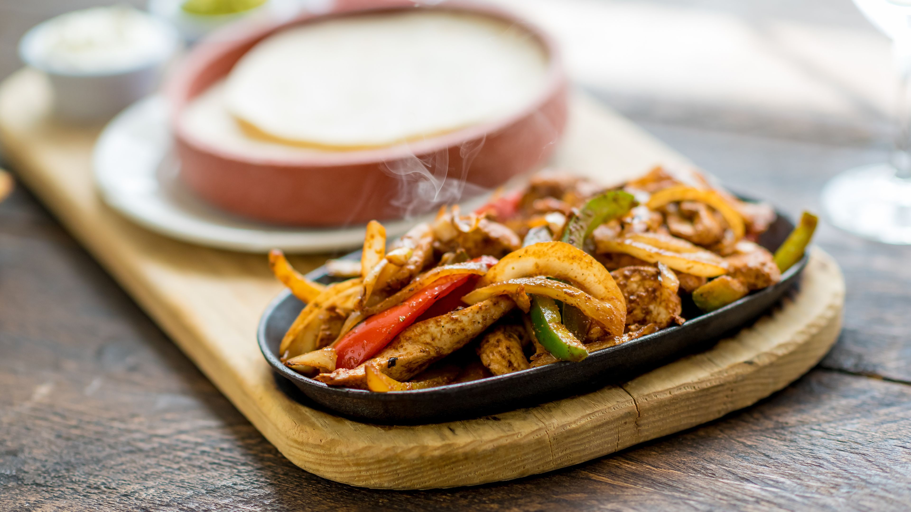 A skillet with fajitas on a wooden board, a bowl of dipping sauce, and a glass of water on a wooden table.