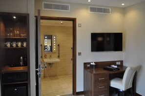 Habitación de hotel con escritorio de madera, televisor montado en la pared, zona de ducha y un armario con utensilios de cocina.