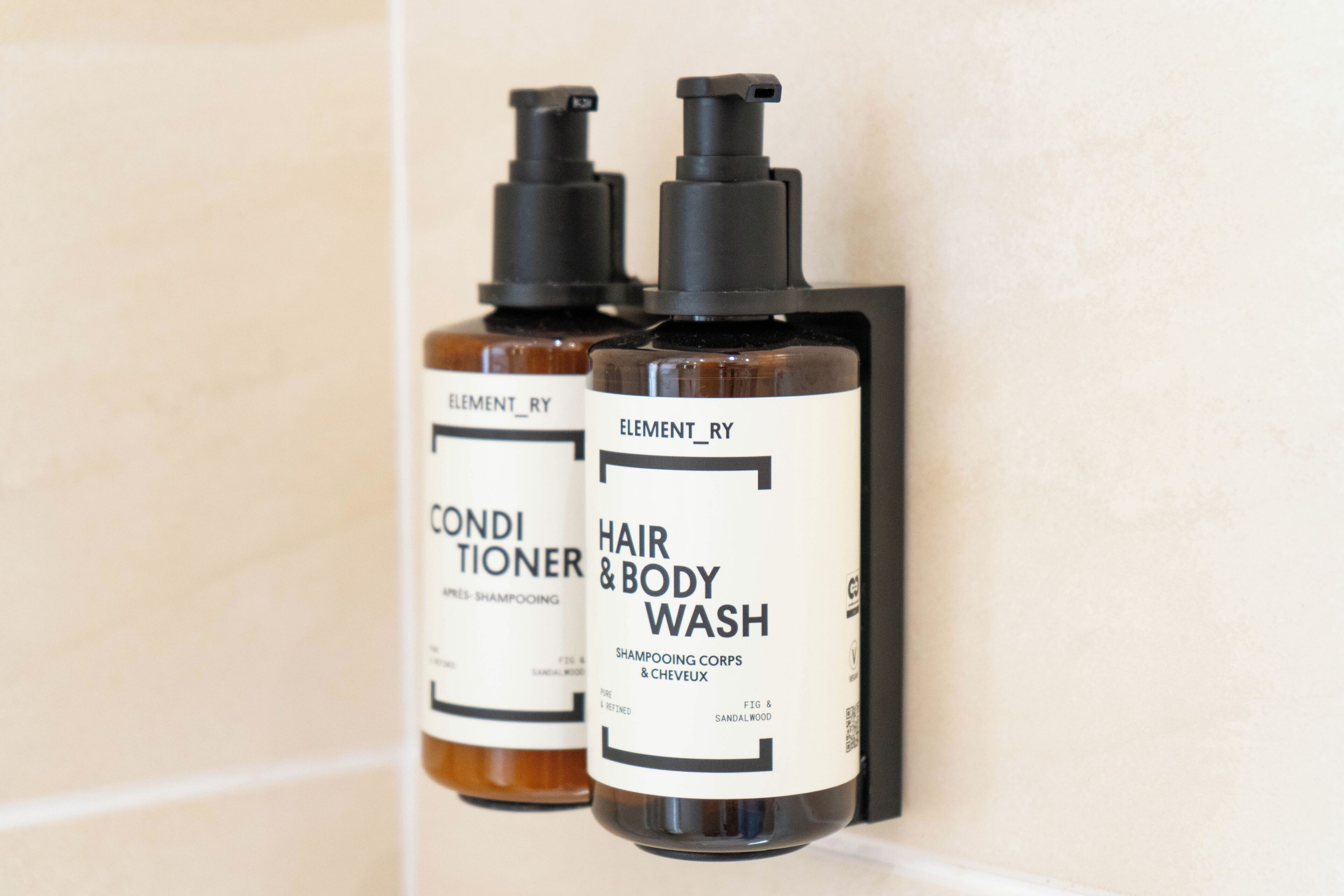 Twee flessen Elementry haar- en bodywash op een betegelde muur.