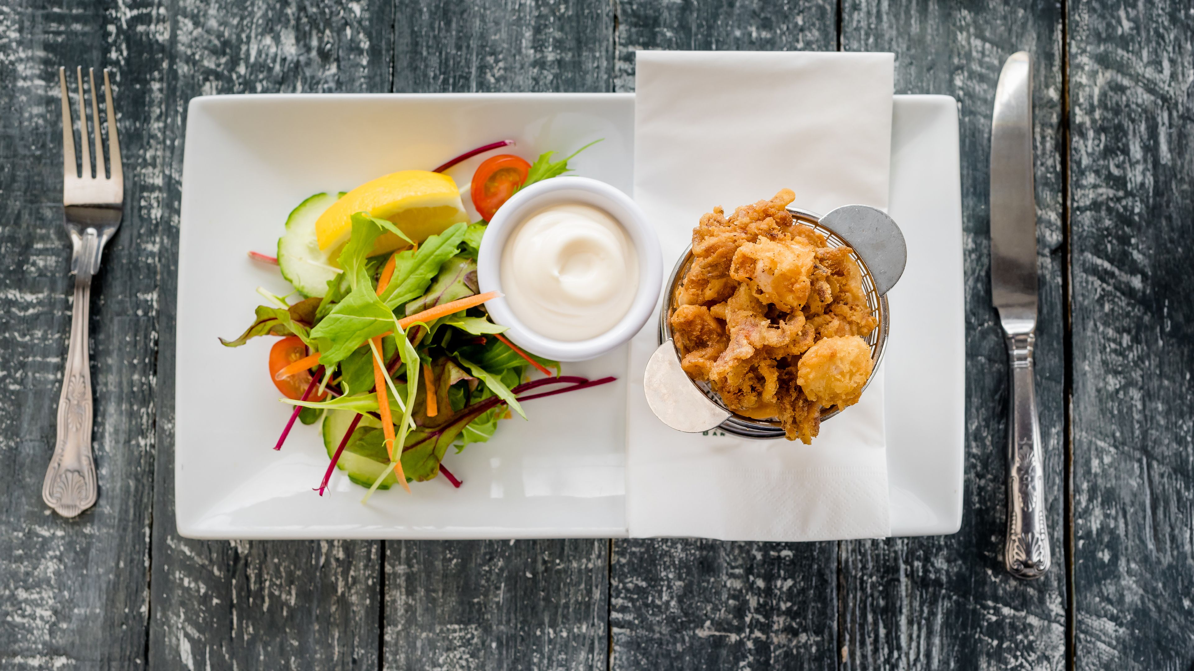 A white rectangular plate with a salad, a bowl of dipping sauce, and a basket of fried food on a wooden table.