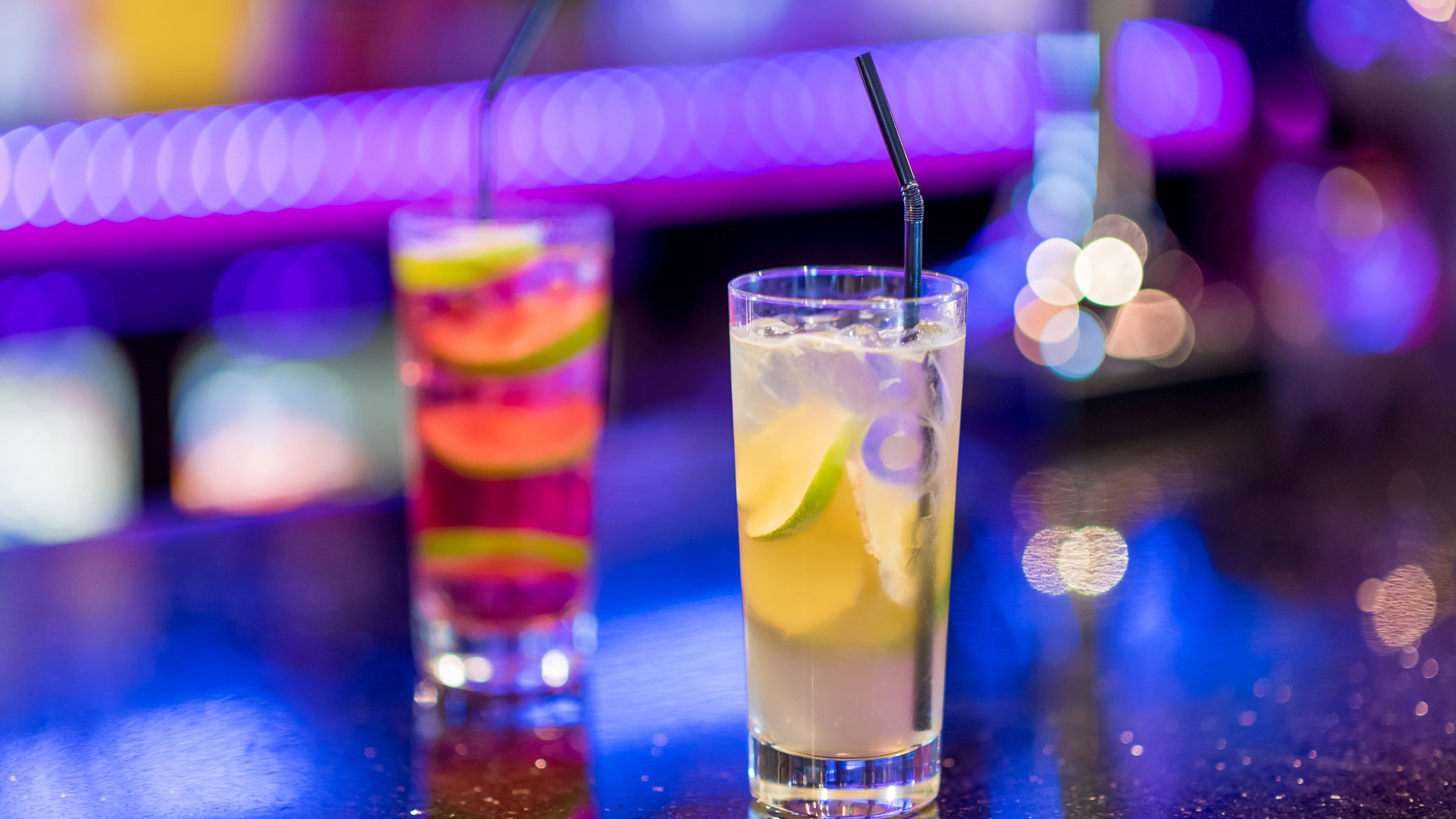 Twee glazen ijskoude drankjes met rietjes en partjes limoen op een bar.