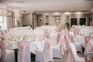 Un salón de banquetes con mesas redondas dispuestas para un evento formal, cada una con manteles blancos y sillas decoradas con cintas rosas.