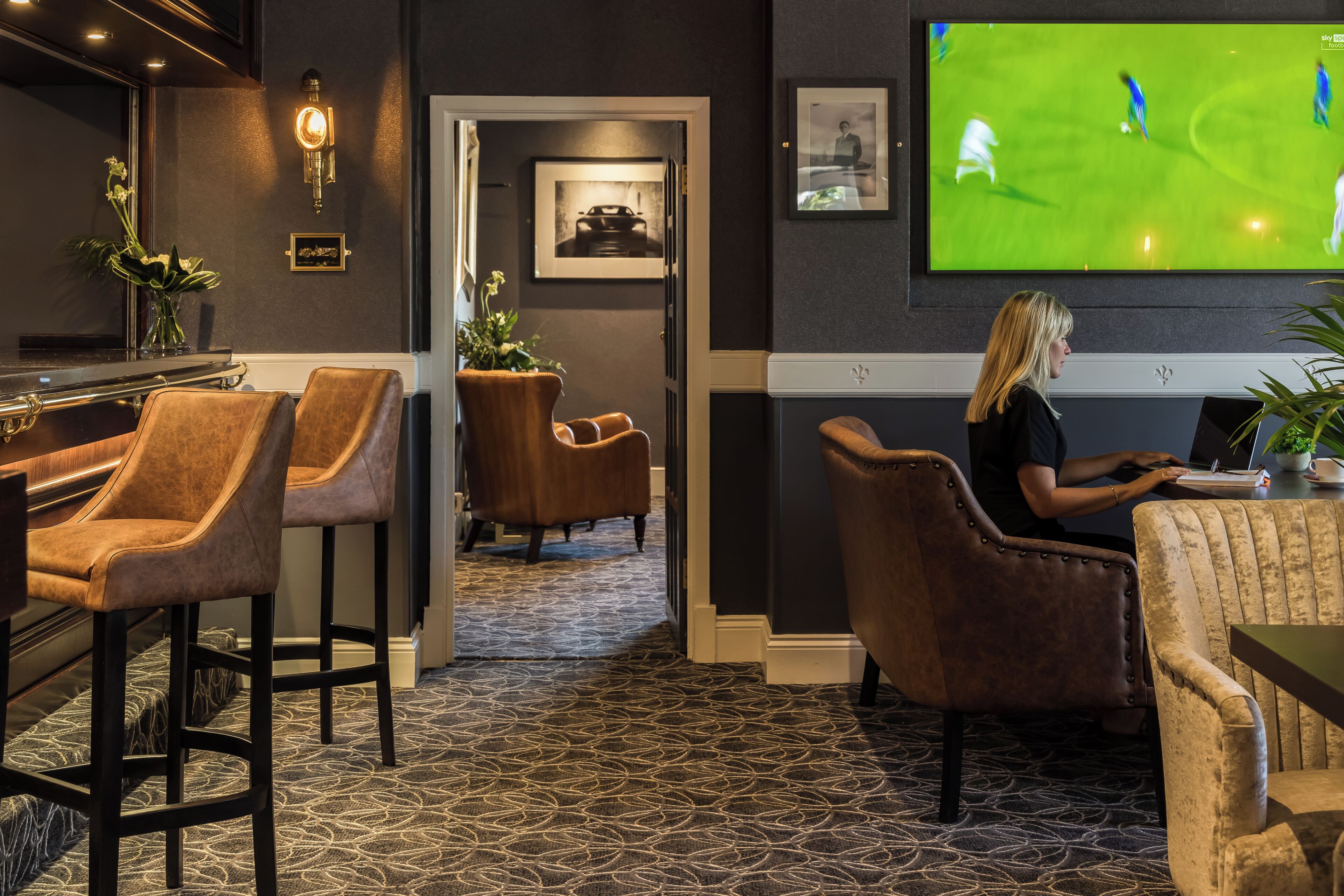 A room with a bar area, seating, a television displaying a sports event, and a door leading to another room.