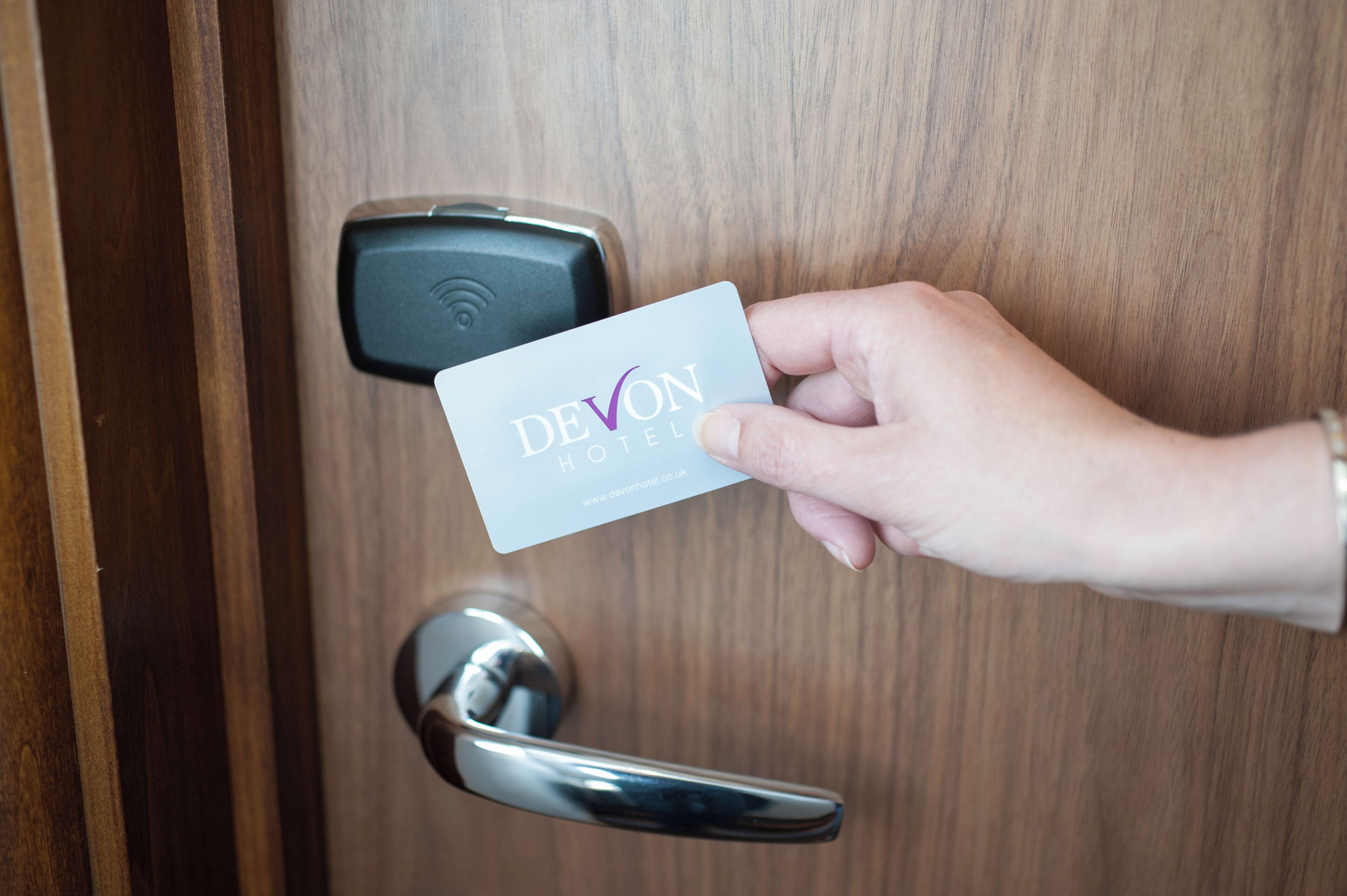 A hand holding a card with 'DEVON HOTEL' written on it, approaching a door with a digital lock.