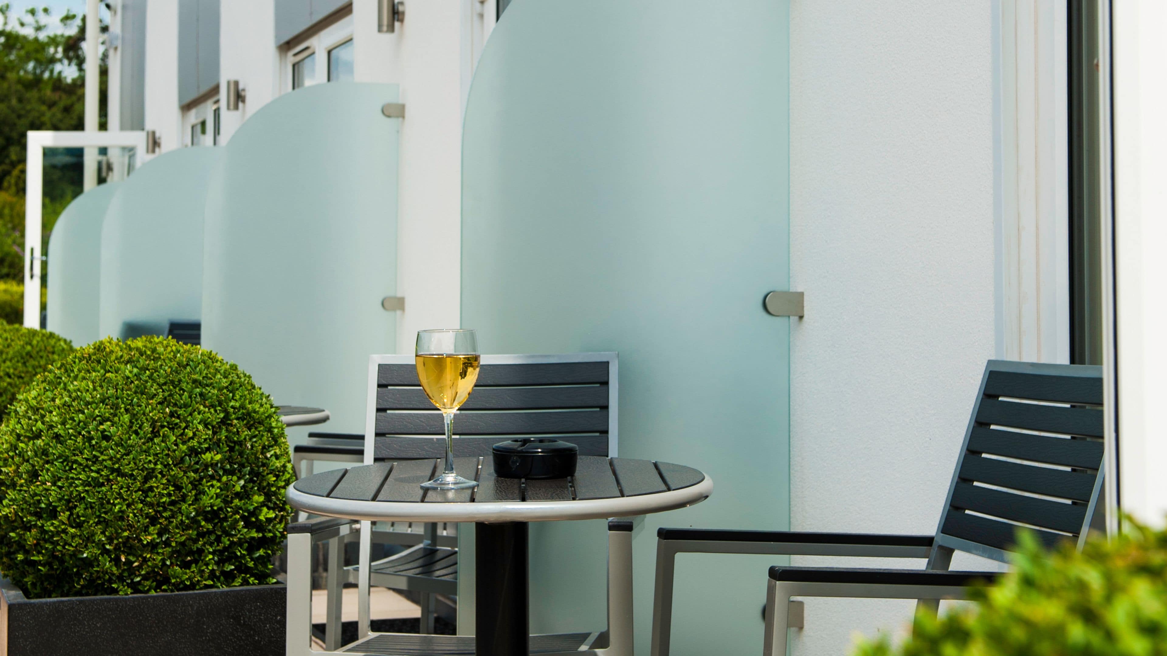 Ein kleiner Sitzbereich im Freien mit Tisch und Stühlen, einem Glas Weißwein und einer ordentlich geschnittenen Hecke.
